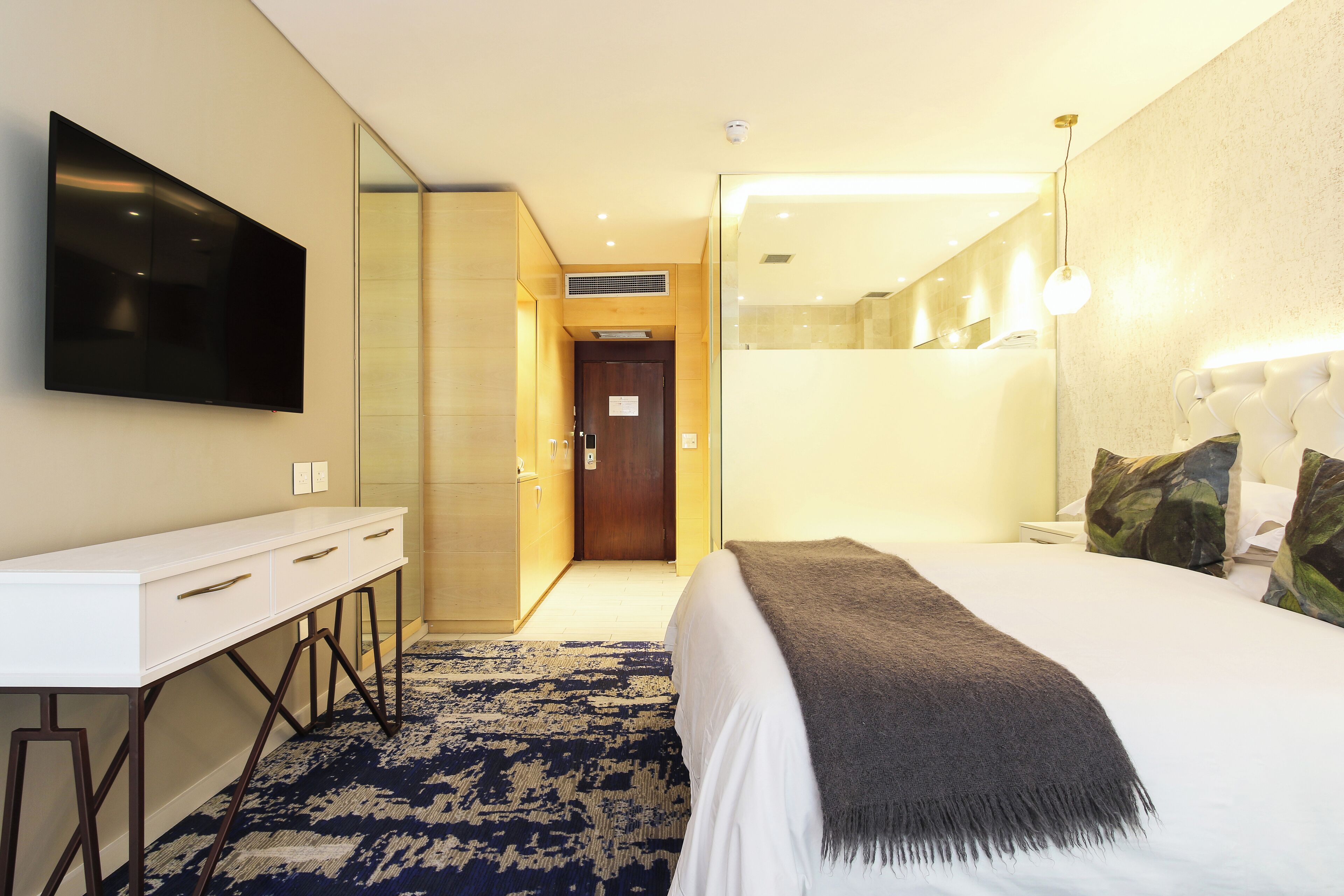 A hotel room with a bed, a TV mounted on the wall, a desk with drawers, a mirror, and a patterned rug.
