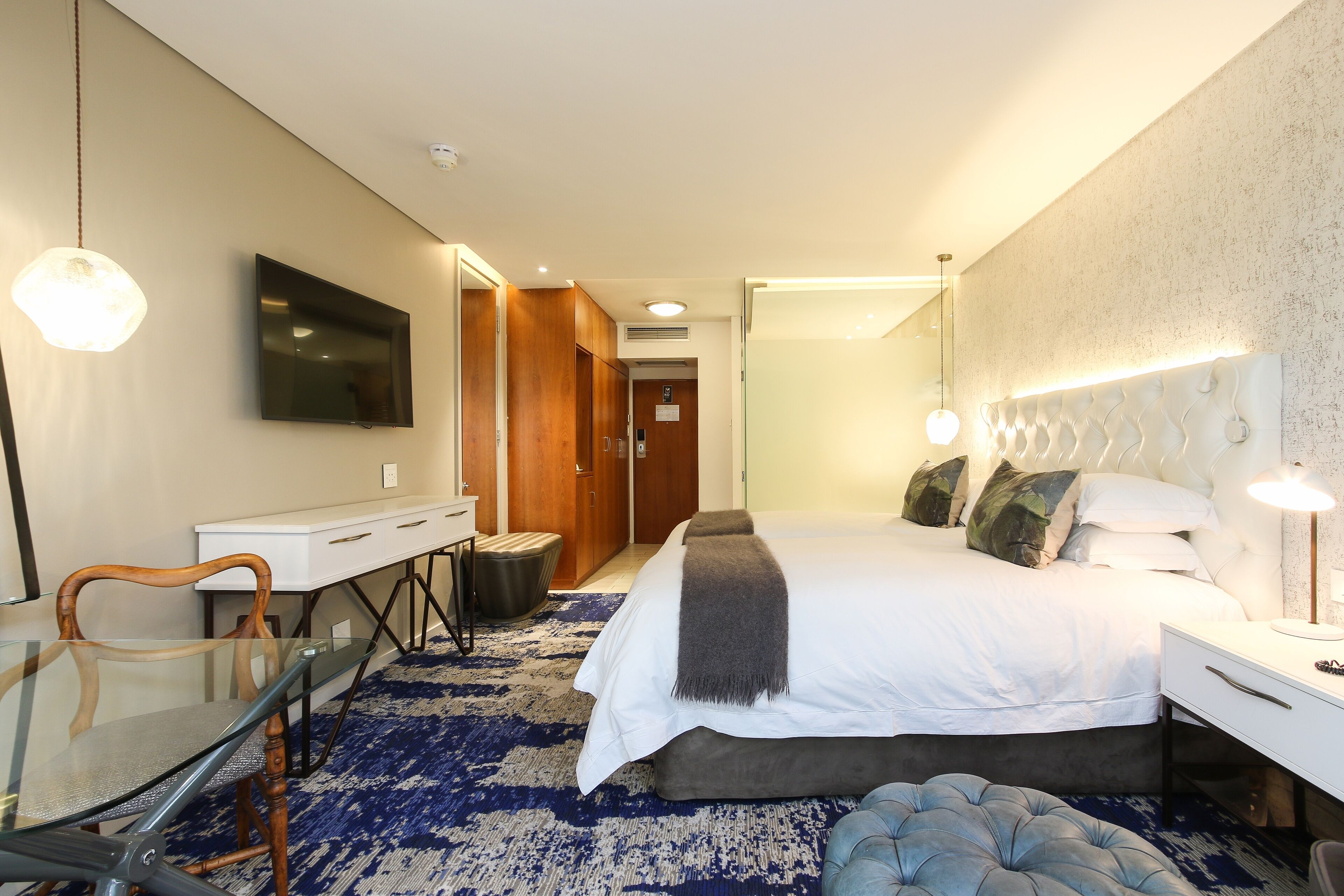 Quarto de hotel moderno com uma cama grande, uma escrivaninha com cadeira, televisão de tela plana e uma mesinha de cabeceira com abajur.