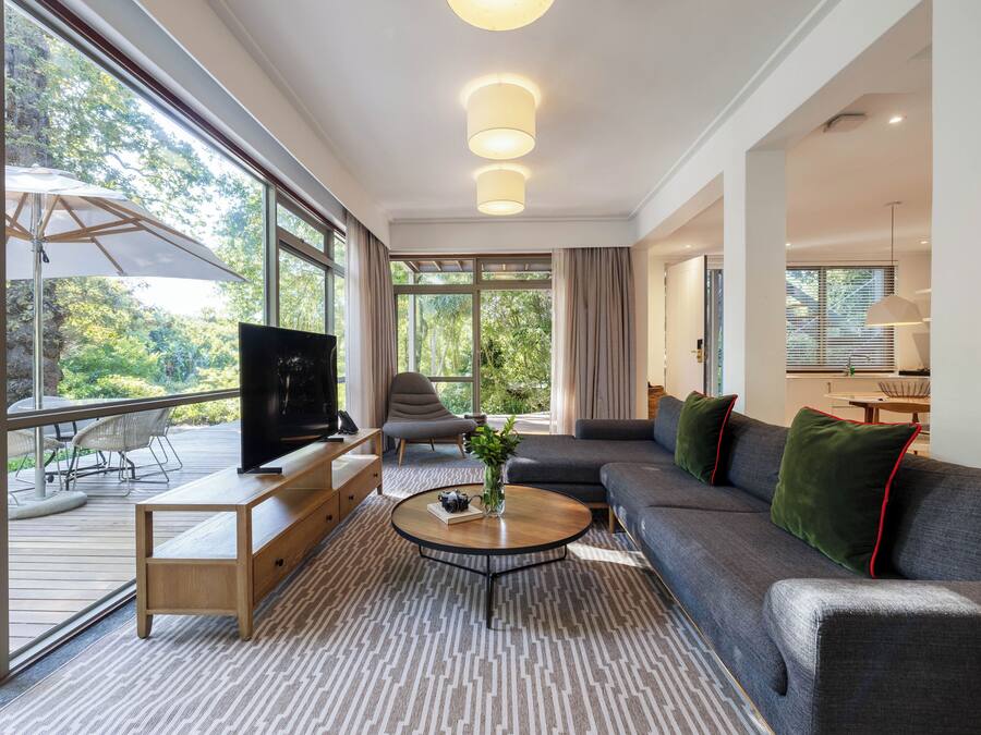 Una sala de estar moderna con un sofá grande, una mesa de centro y un televisor sobre un mueble de madera. Hay un balcón con mobiliario exterior y vistas a zonas verdes.