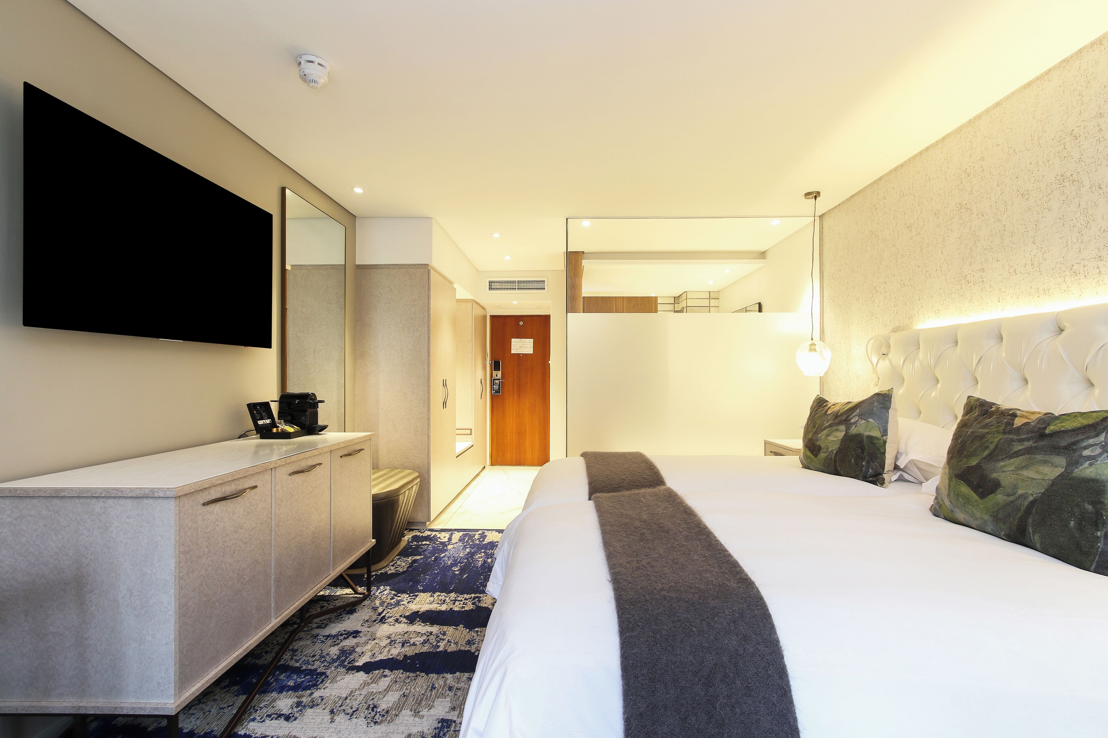 Ein Hotelzimmer mit einem großen Bett, einem flachbildfernseher an der Wand, einem Nachttisch mit Lampe und einem durchgehenden Türspalt einsehbaren Badezimmer.