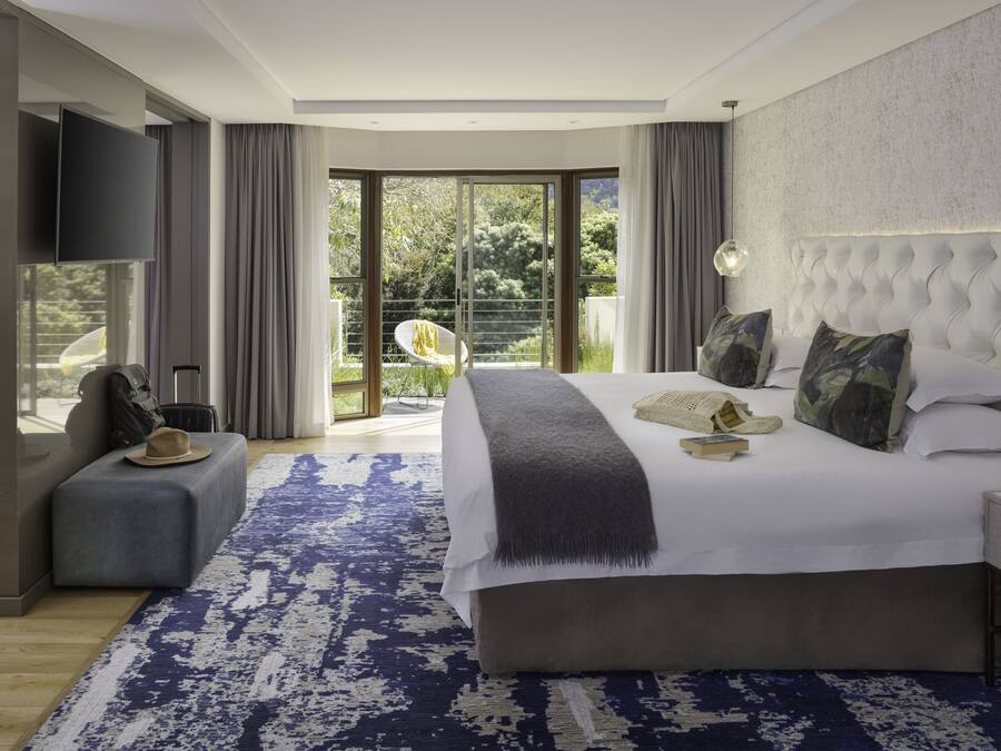Una habitación de hotel moderna con una cama grande, un televisor de pantalla plana, una zona de estar con una silla y un balcón con vistas a zonas verdes.