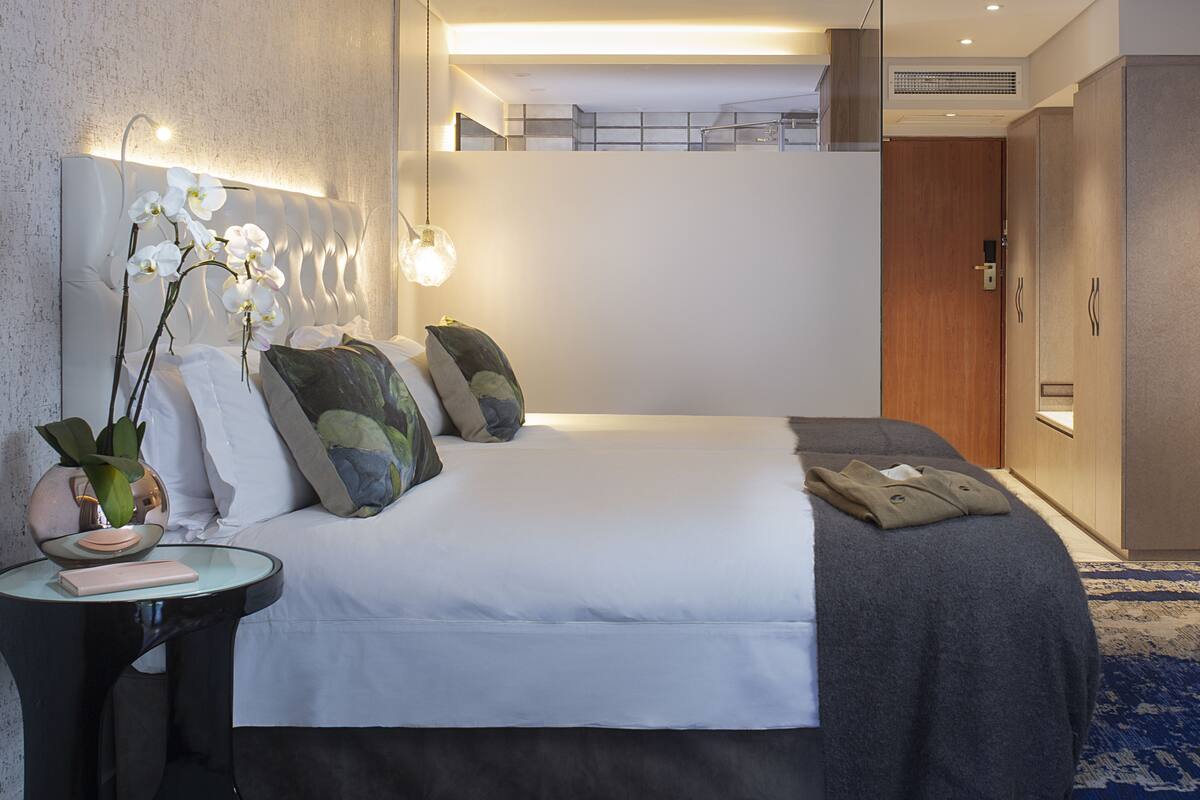A modern hotel room with a large bed, a bedside table with a lamp, a small side table, and a blue patterned carpet.