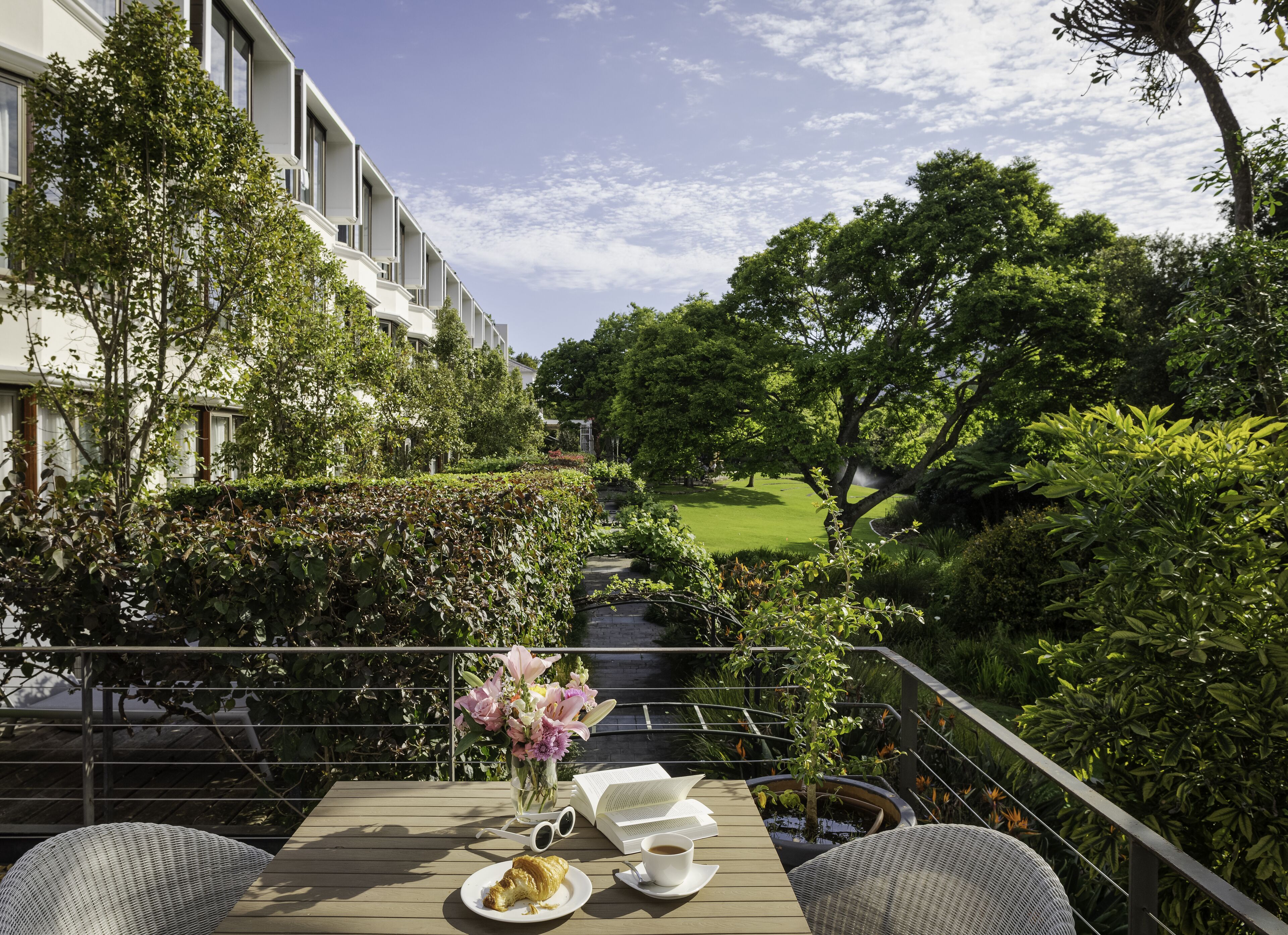 A terrace with a table set for a meal, surrounded by lush greenery and a view of a well-maintained lawn and trees.