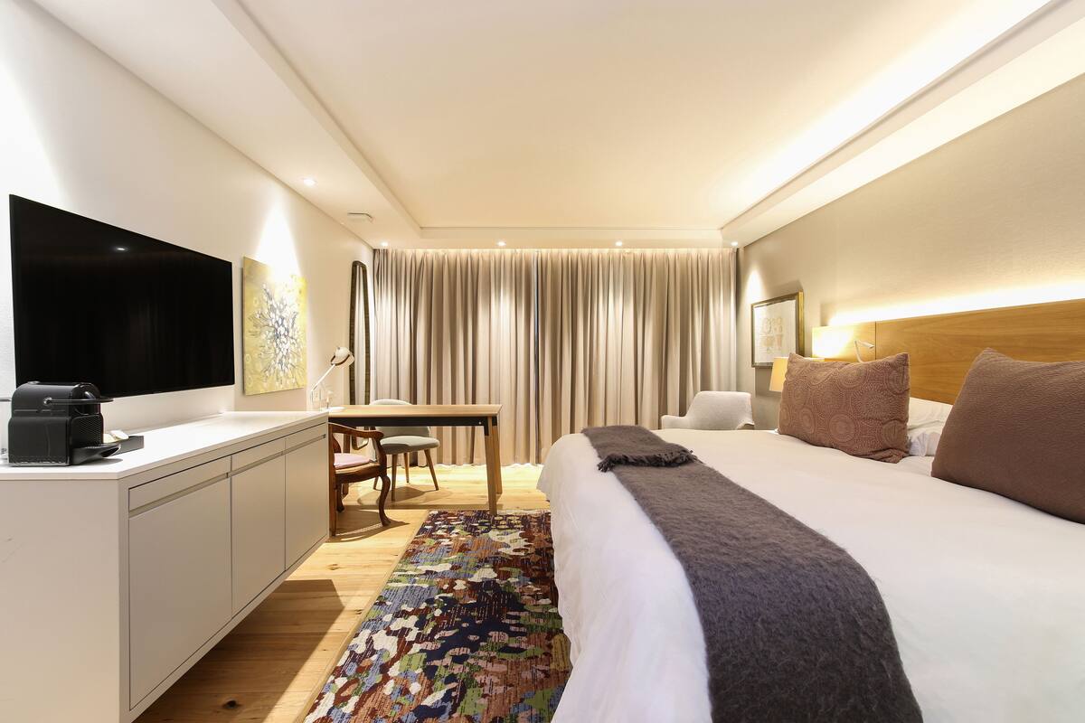 A modern hotel room with a large bed, a desk, a flat-screen TV, and a large window with curtains.