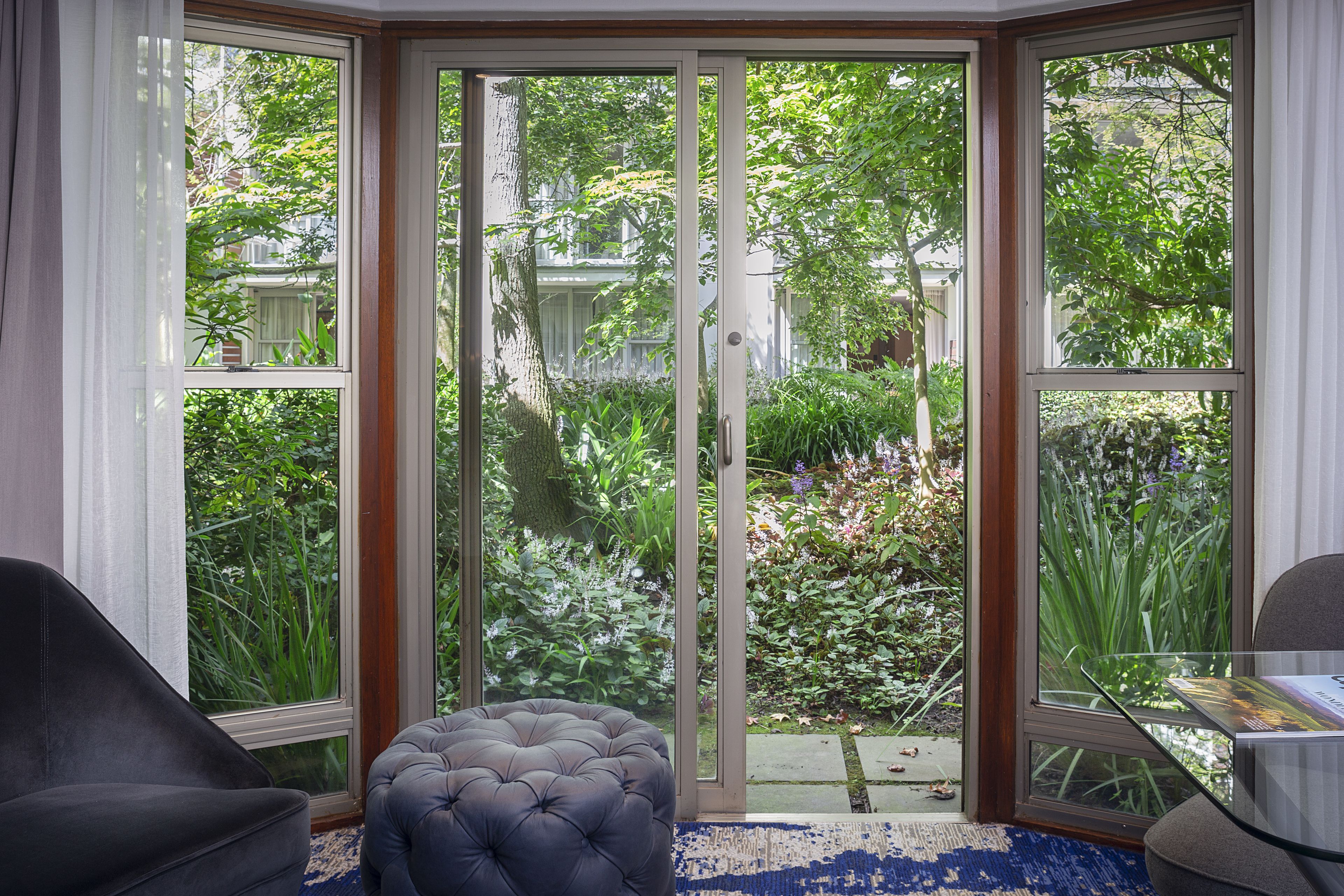 A room with a sliding glass door leading to a garden, a tufted ottoman, and a glass table with a book.