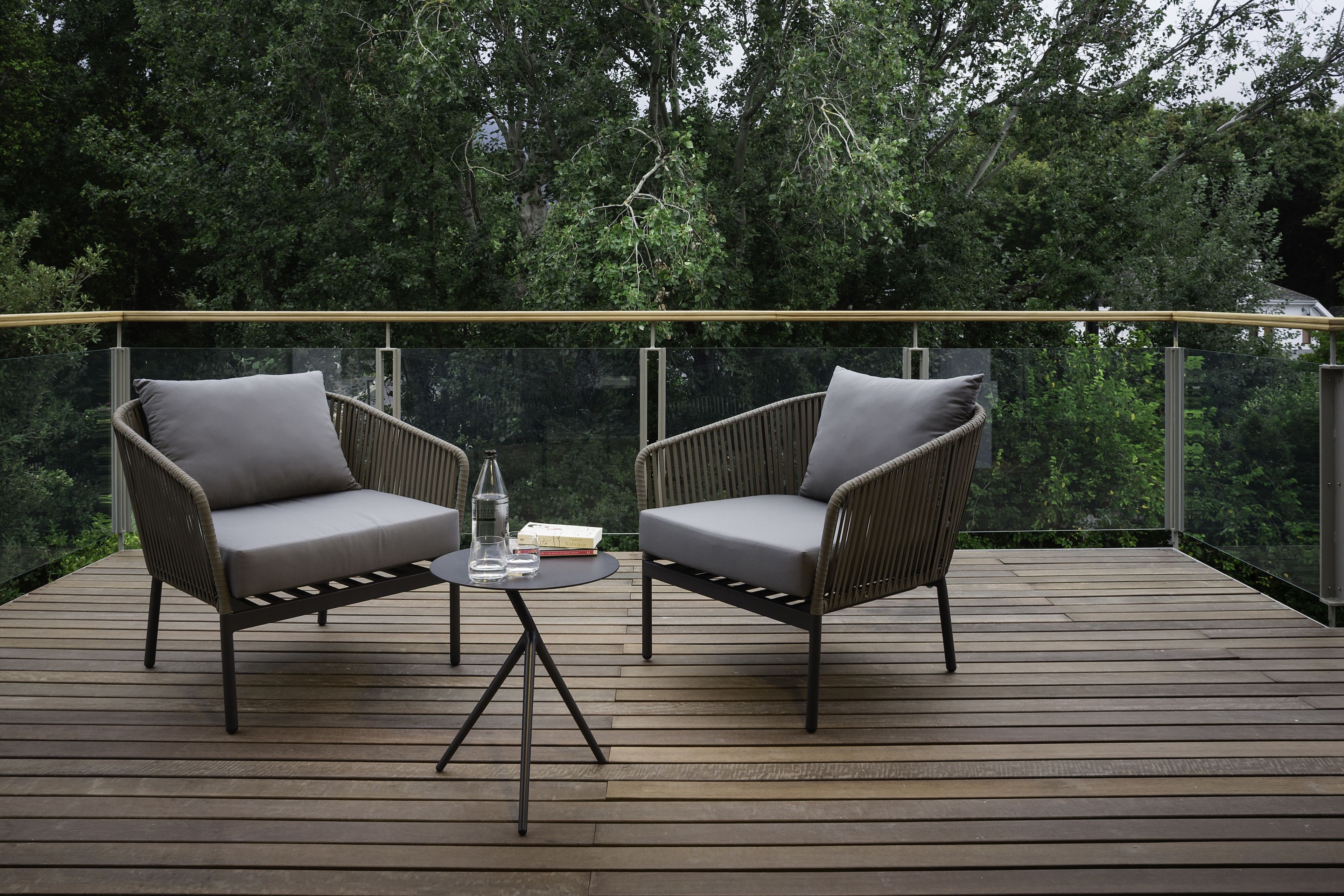 A wooden deck with two wicker chairs and a small table, surrounded by a glass balustrade and dense greenery.