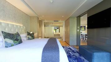 A modern hotel room with a large bed, a blue bench, a flat-screen TV, and a bathroom visible through a glass door.