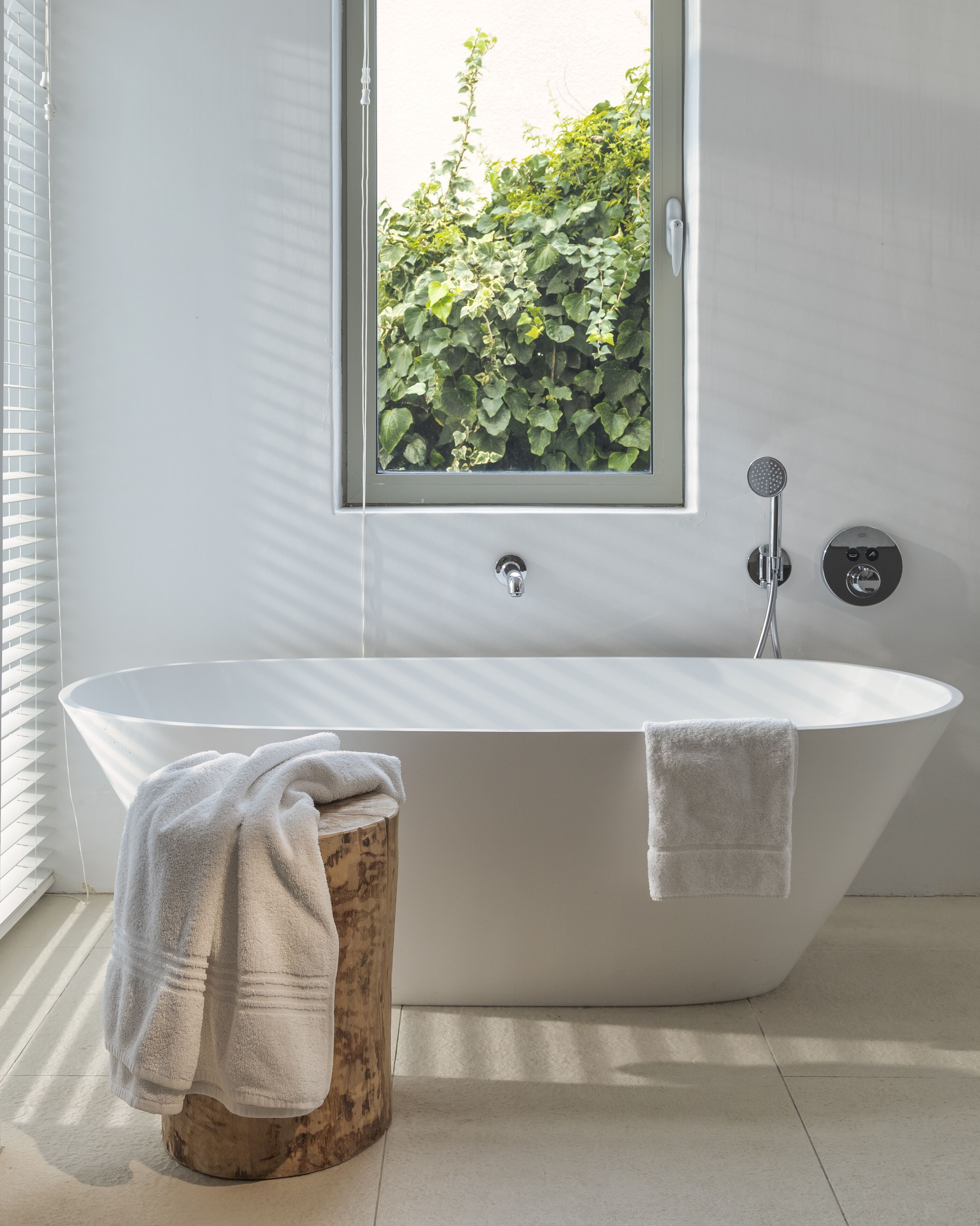 Un baño moderno con una bañera independiente, un taburete de madera y un ventanal con vistas a la vegetación.