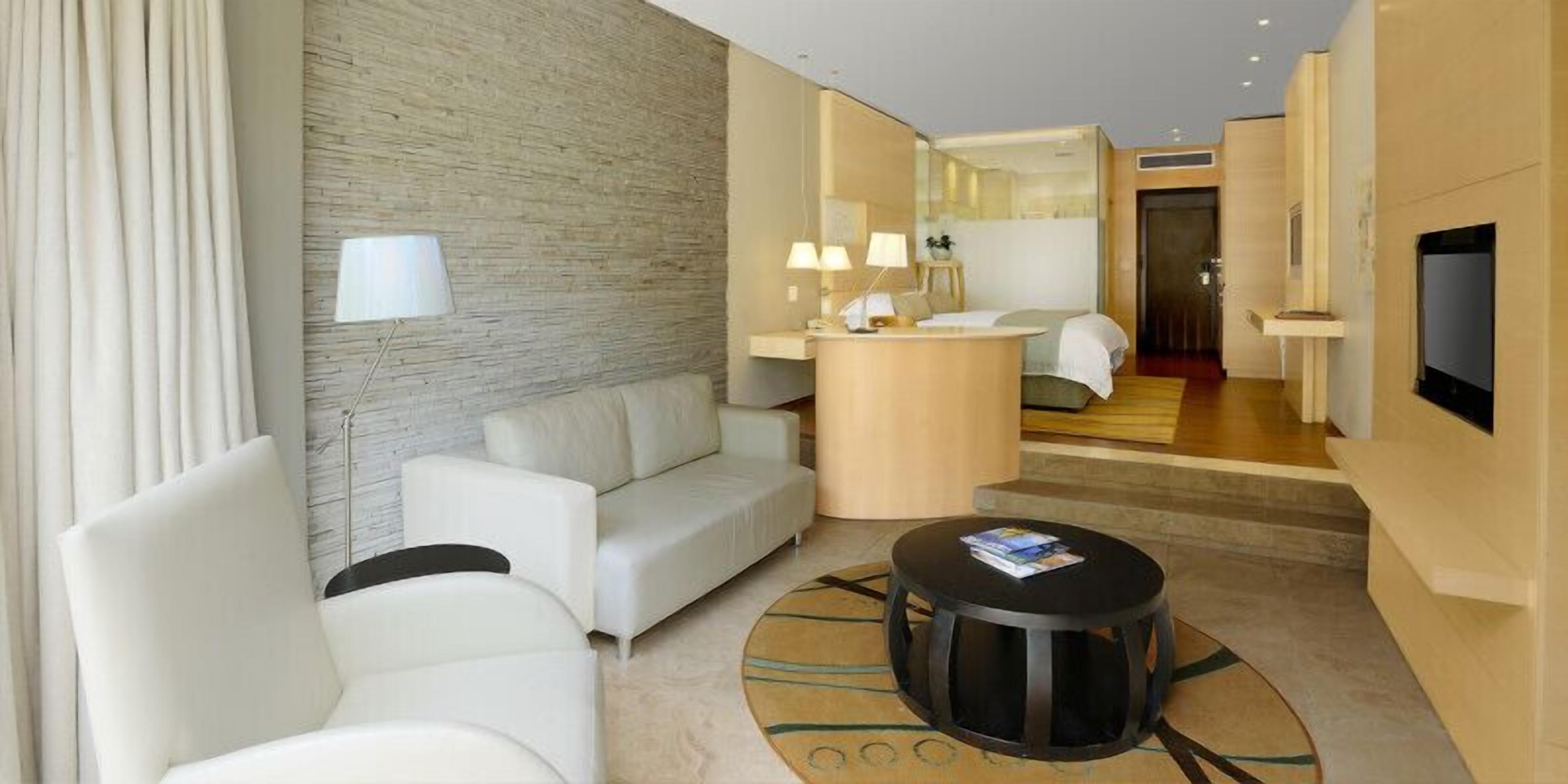 Une chambre d’hôtel moderne, équipée d’un canapé, d’une table basse ronde, d’un petit bureau avec des lampes et d’un téléviseur.
