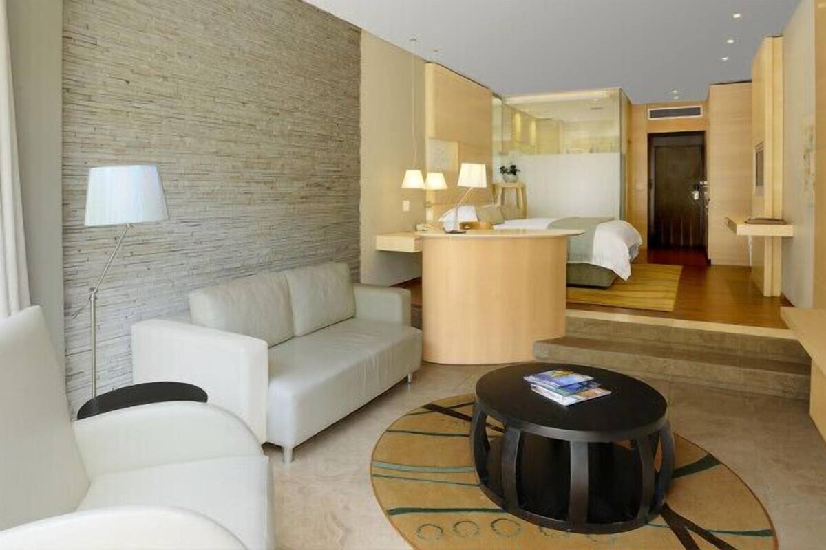 Une chambre d’hôtel moderne, équipée d’un canapé, d’une table basse ronde, d’un petit bureau avec des lampes et d’un téléviseur.