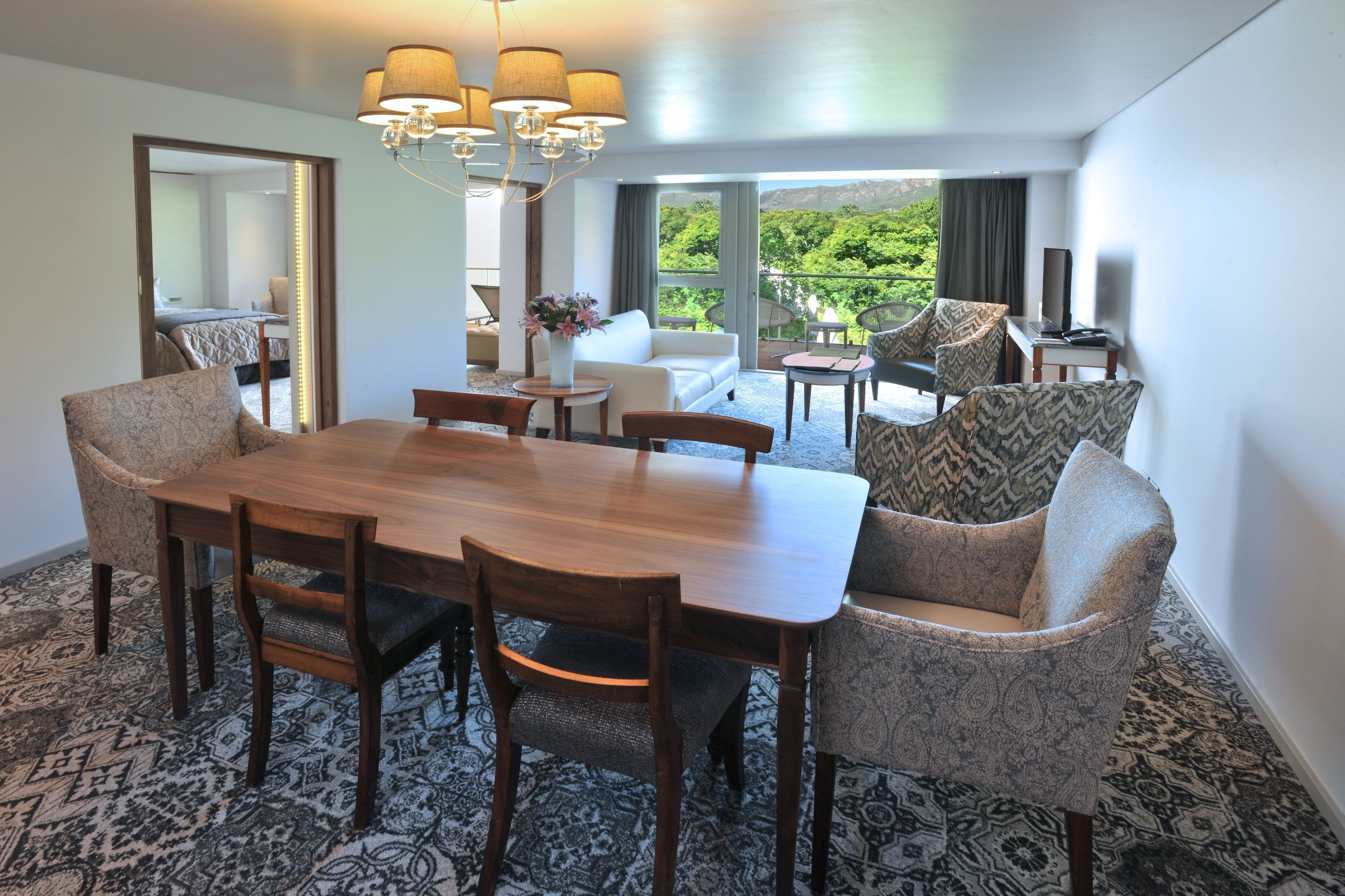 A modern hotel room with a dining area featuring a wooden table and patterned chairs, a sofa, and a balcony with a view of greenery.