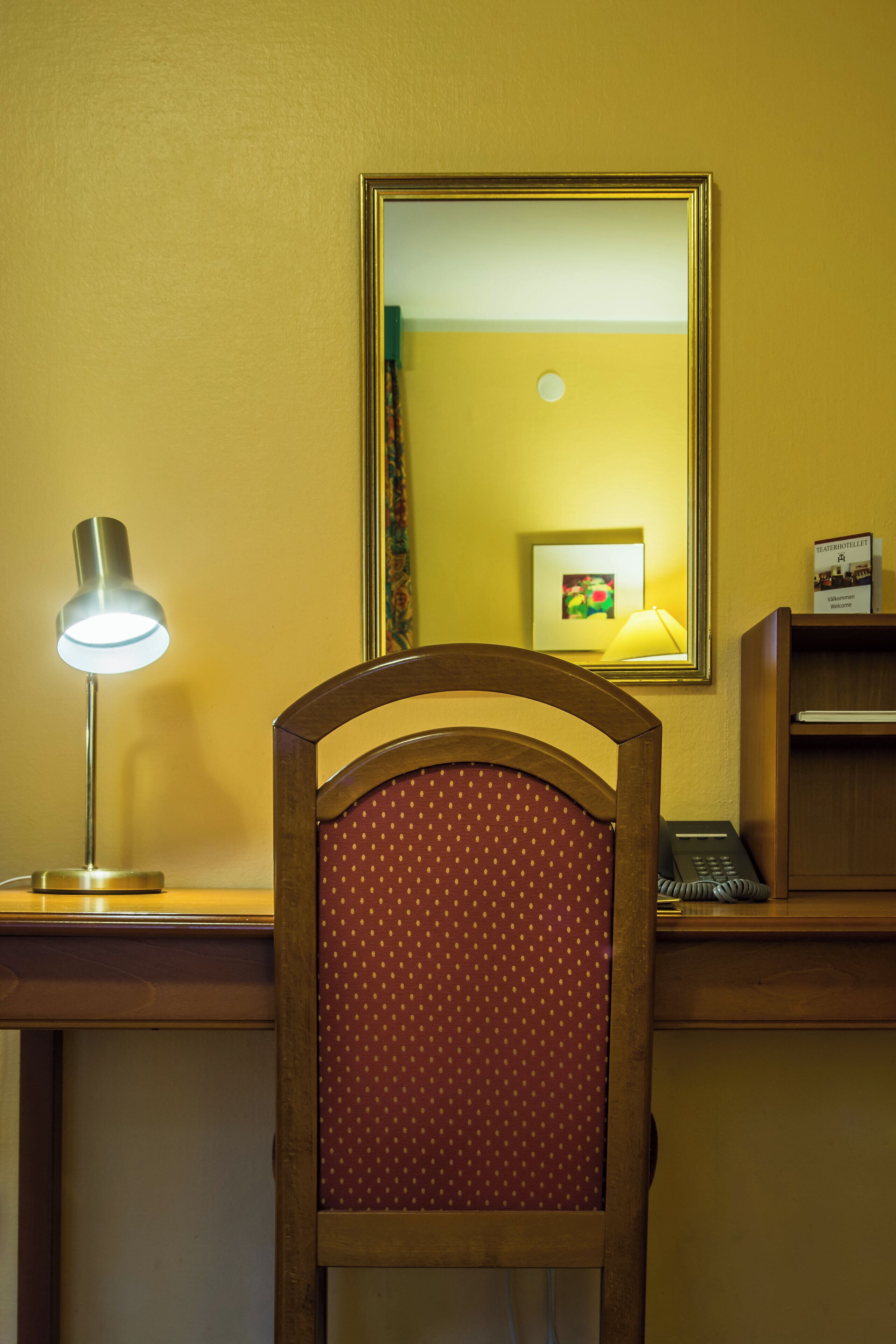 Habitación de hotel con escritorio de madera, lámpara de escritorio, teléfono y espejo que refleja un cuadro enmarco y una lámpara.