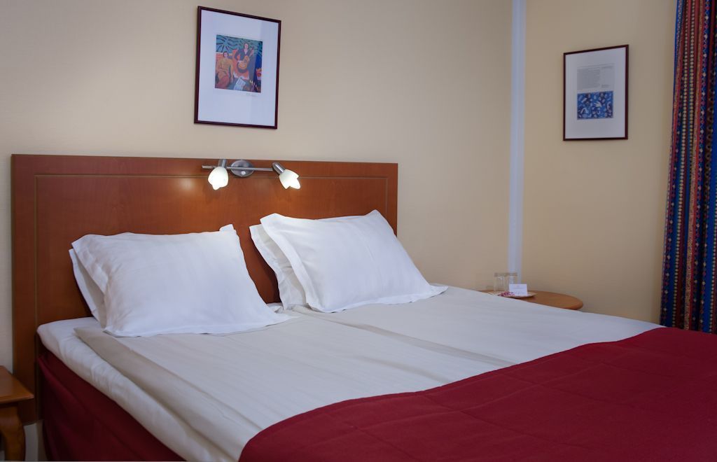 A hotel room with a wooden headboard, two white pillows, a red bedspread, and two bedside tables with lamps.