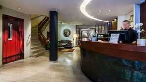 A hotel lobby with a reception desk, a staircase, a red curtain, and a wall with a circular light fixture. - Teaterhotellet (Malmö)