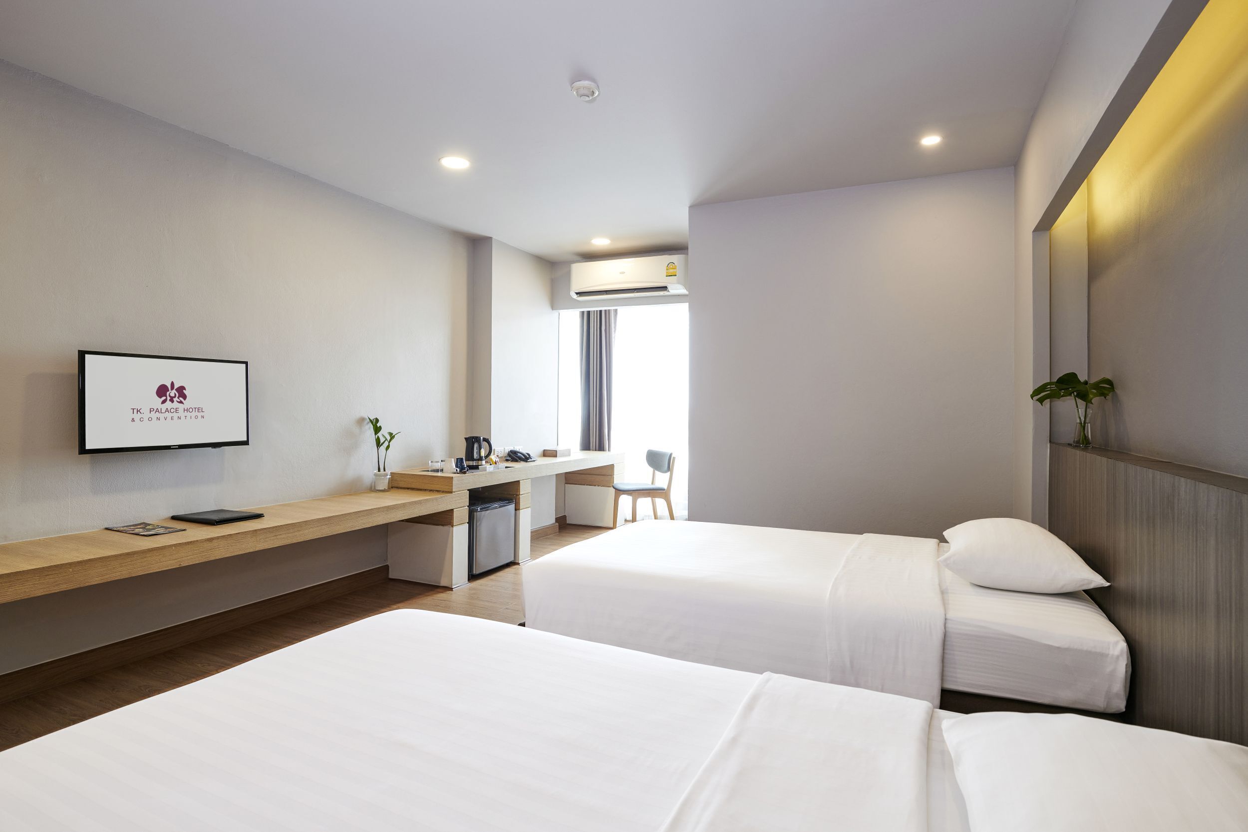 A hotel room with two beds, a desk with a chair, a small kitchenette, and a TV mounted on the wall.