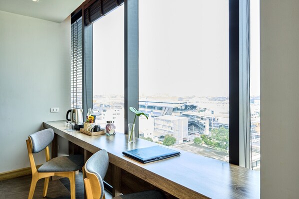 Una habitación moderna con un escritorio de madera, dos sillas, una computadora portátil, un florero con una planta y vistas a un paisaje urbano a través de amplios ventanales.