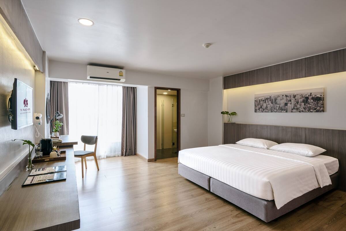 A modern hotel room with a large bed, a desk with a chair, a TV, and a cityscape artwork on the wall.