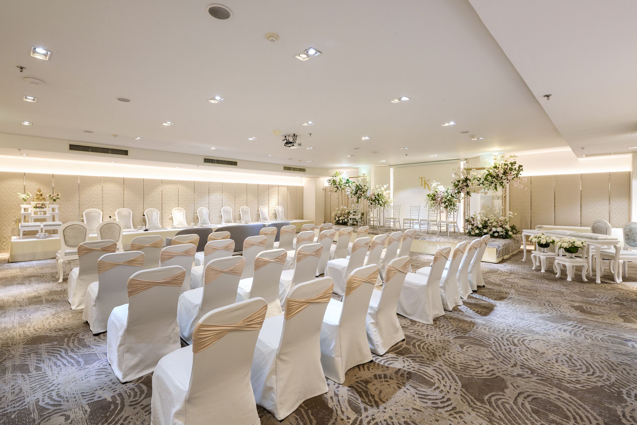 A large conference room with rows of white chairs, a long table, and floral decorations.