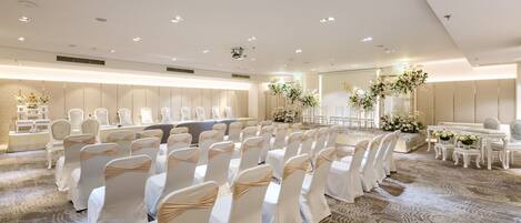 A large conference room with rows of white chairs, a long table, and floral decorations.