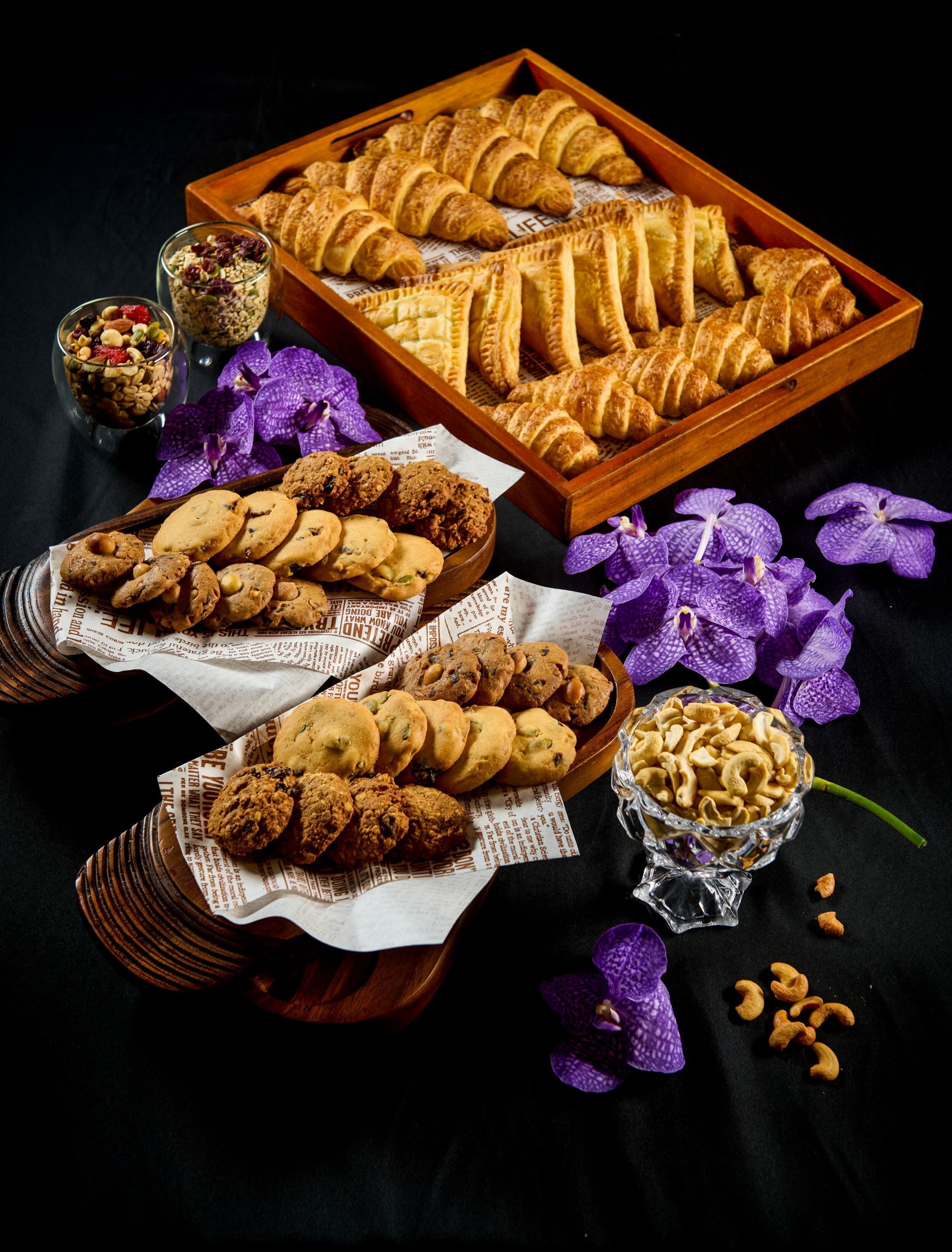 Diversas opciones de panadería, incluyendo cruasanes, galletas y pasteles, dispuestas en bandejas y tazones, con orquídeas moradas y un fondo oscuro.