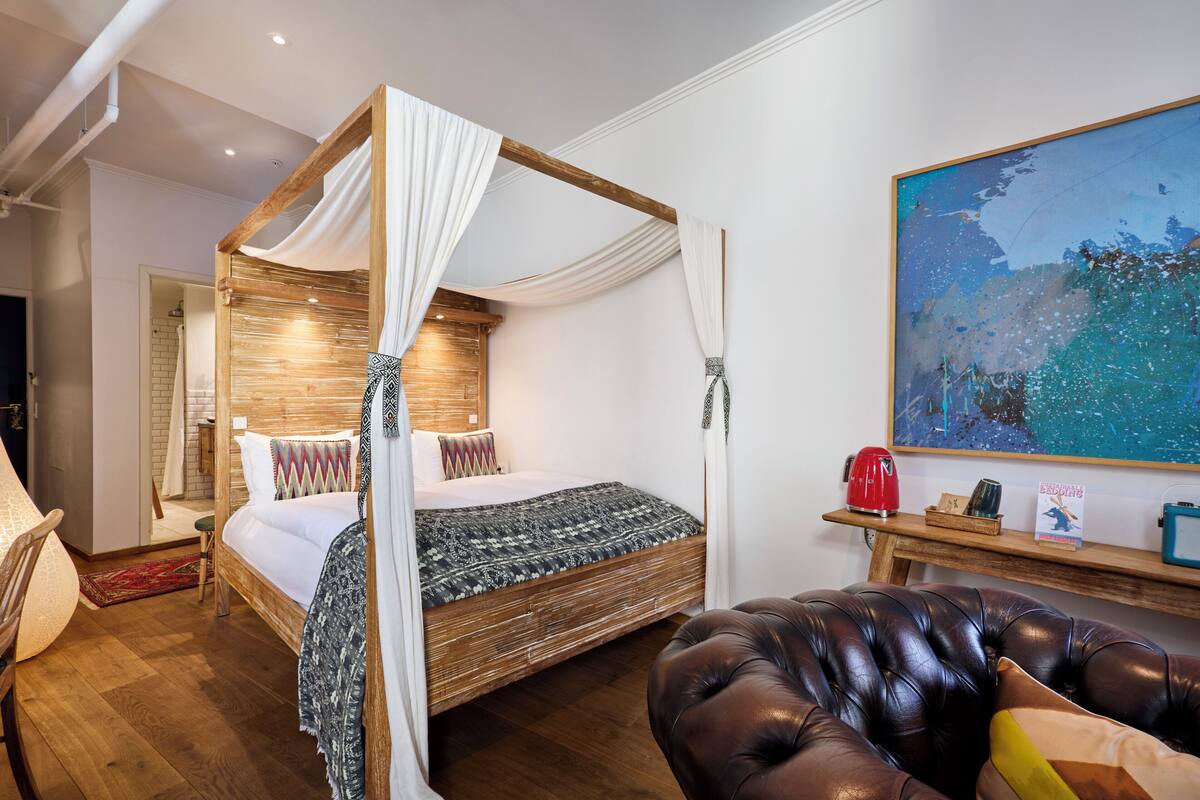 A four-poster bed with a canopy, a wooden headboard, a patterned bedspread, a red kettle, a bookshelf with books and a picture frame, and a large abstract painting on the wall.