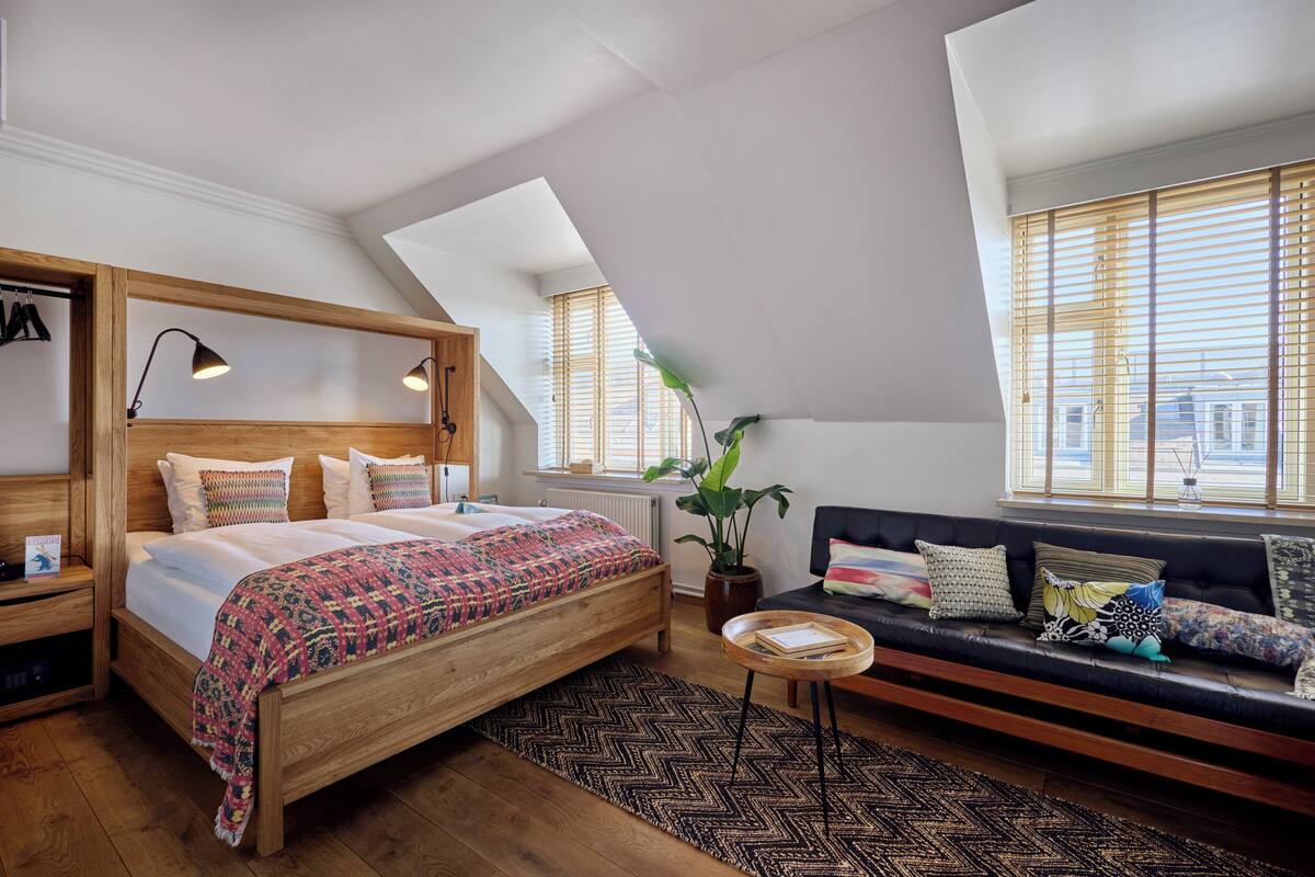 A bedroom with a wooden bed, a black sofa, a small round table, and a large window with blinds.