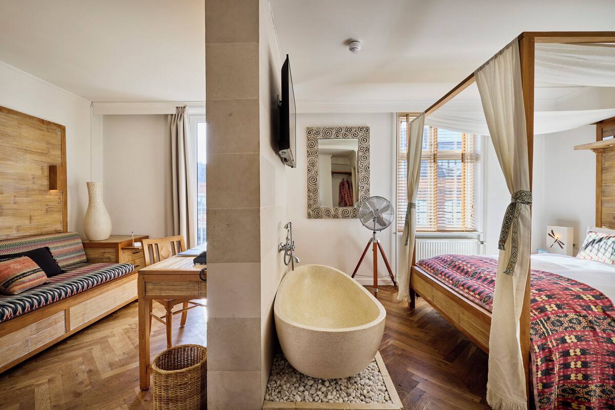 A bedroom with a four-poster bed, a sitting area with a sofa, a bathtub, and a television mounted on the wall.