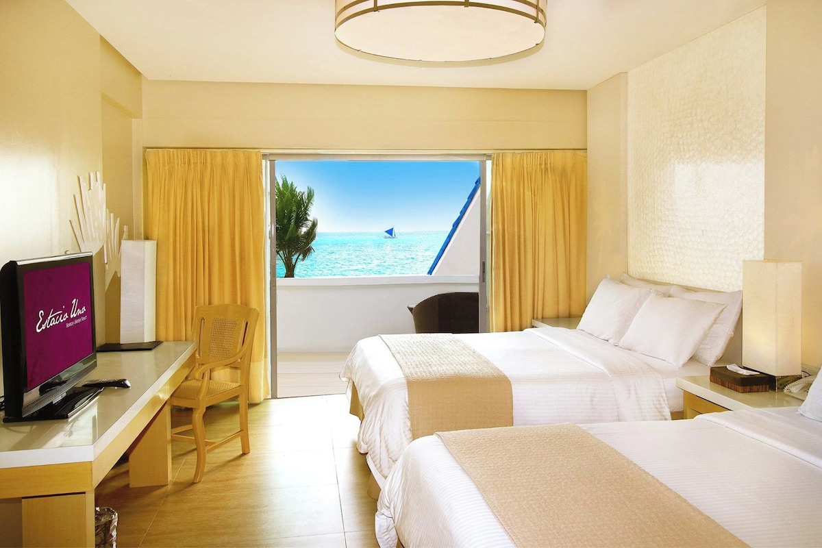 Een hotelkamer met twee bedden, een bureau met een televisie, een stoel en uitzicht op zee.