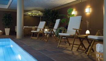 Un área junto a la piscina con sillas de descanso y una pequeña mesa, rodeada de plantas en macetas y un suelo de baldosas.
