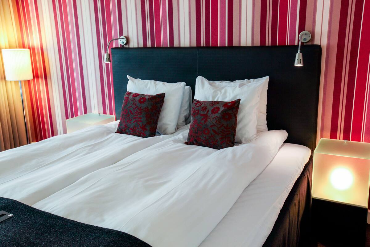 Habitación de hotel con cama, dos almohadas, mesita de noche, lámpara y pared a rayas.
