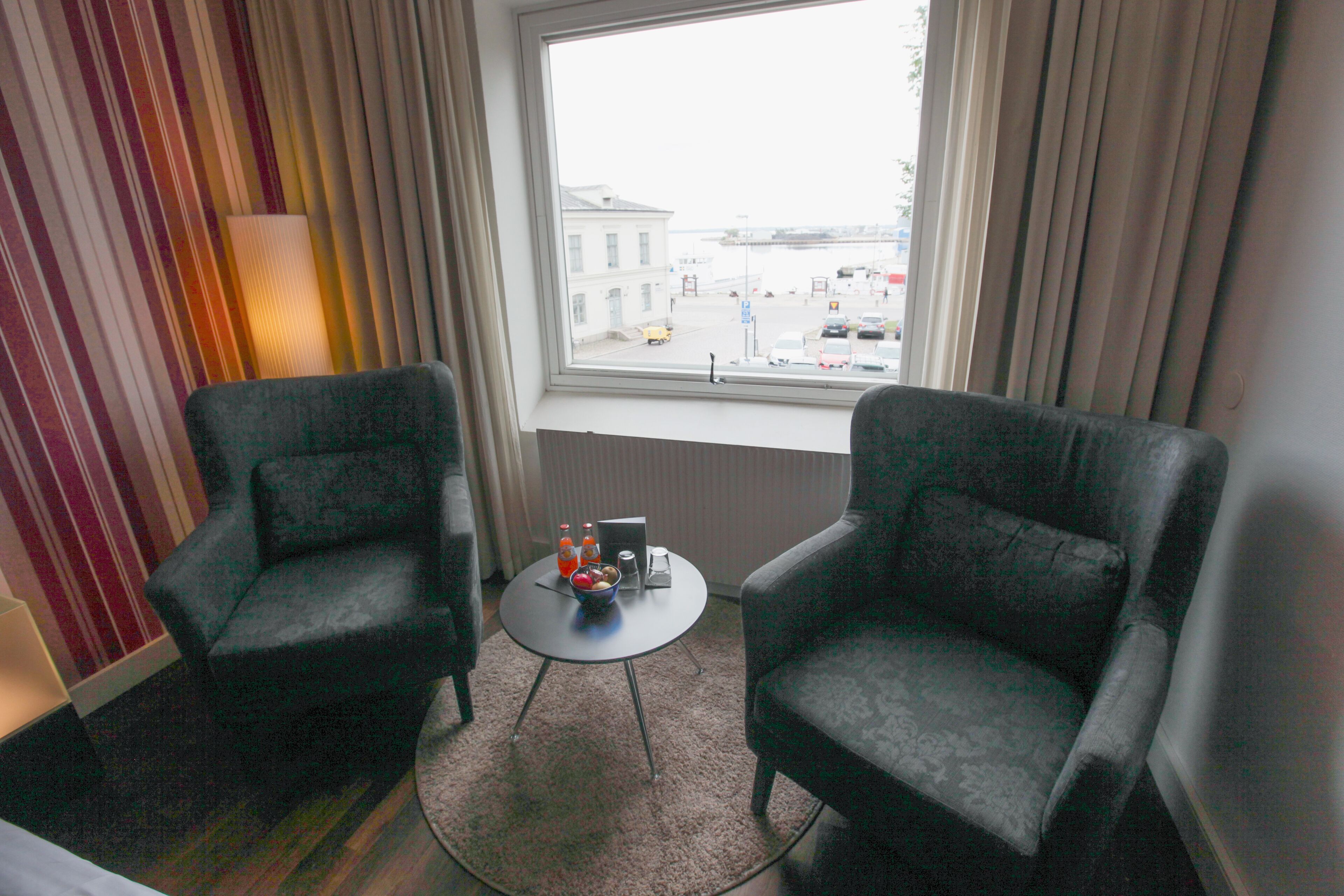 Una habitación con dos sillones, una mesita redonda con un frutero y un ordenador portátil, y vista a una calle a través de una ventana con cortinas.