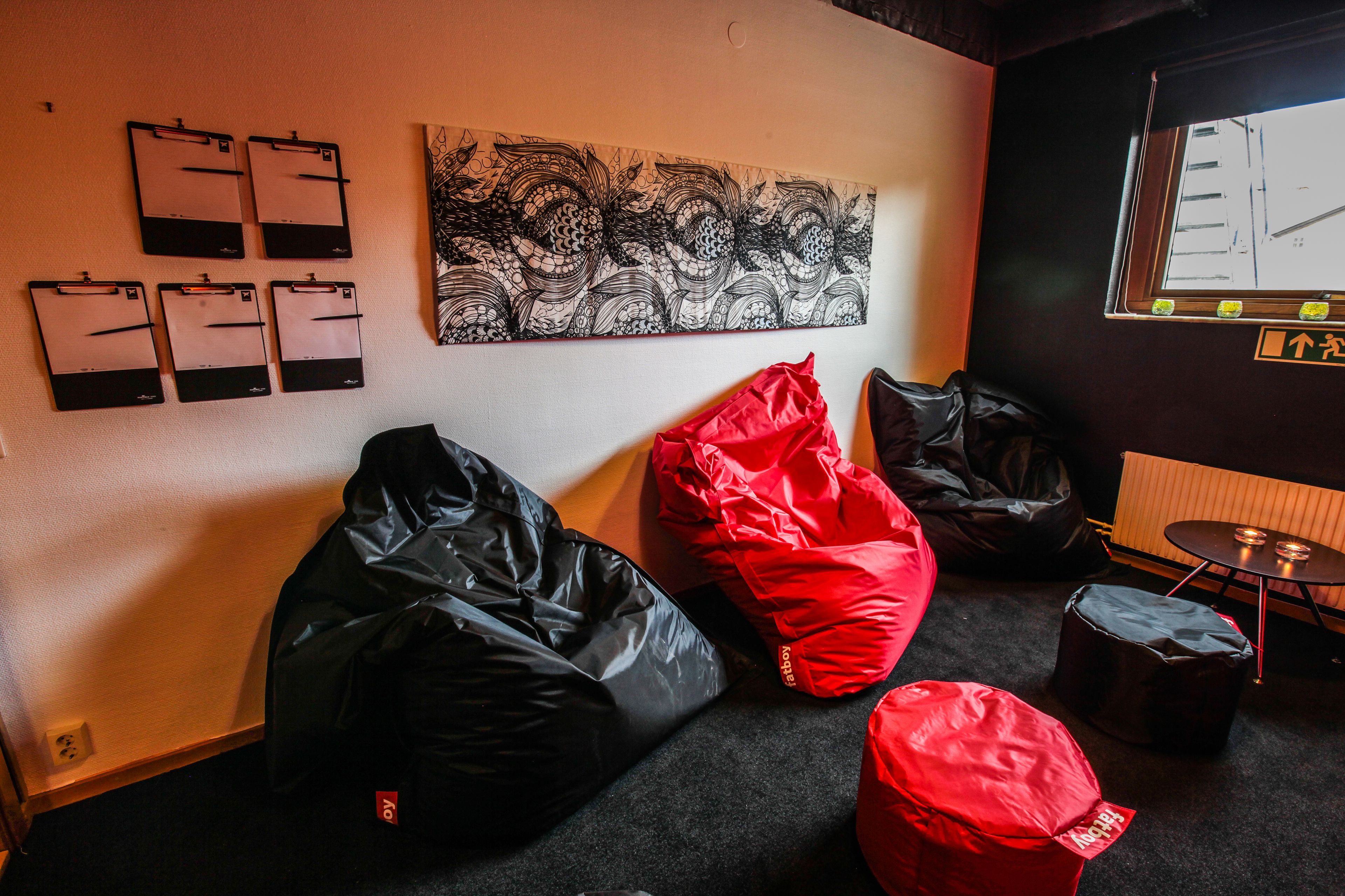 Una habitación con un sillón de saco negro y rojo, otro sillón de saco negro, una obra de arte colgada en la pared, y una ventana con vistas a edificios.