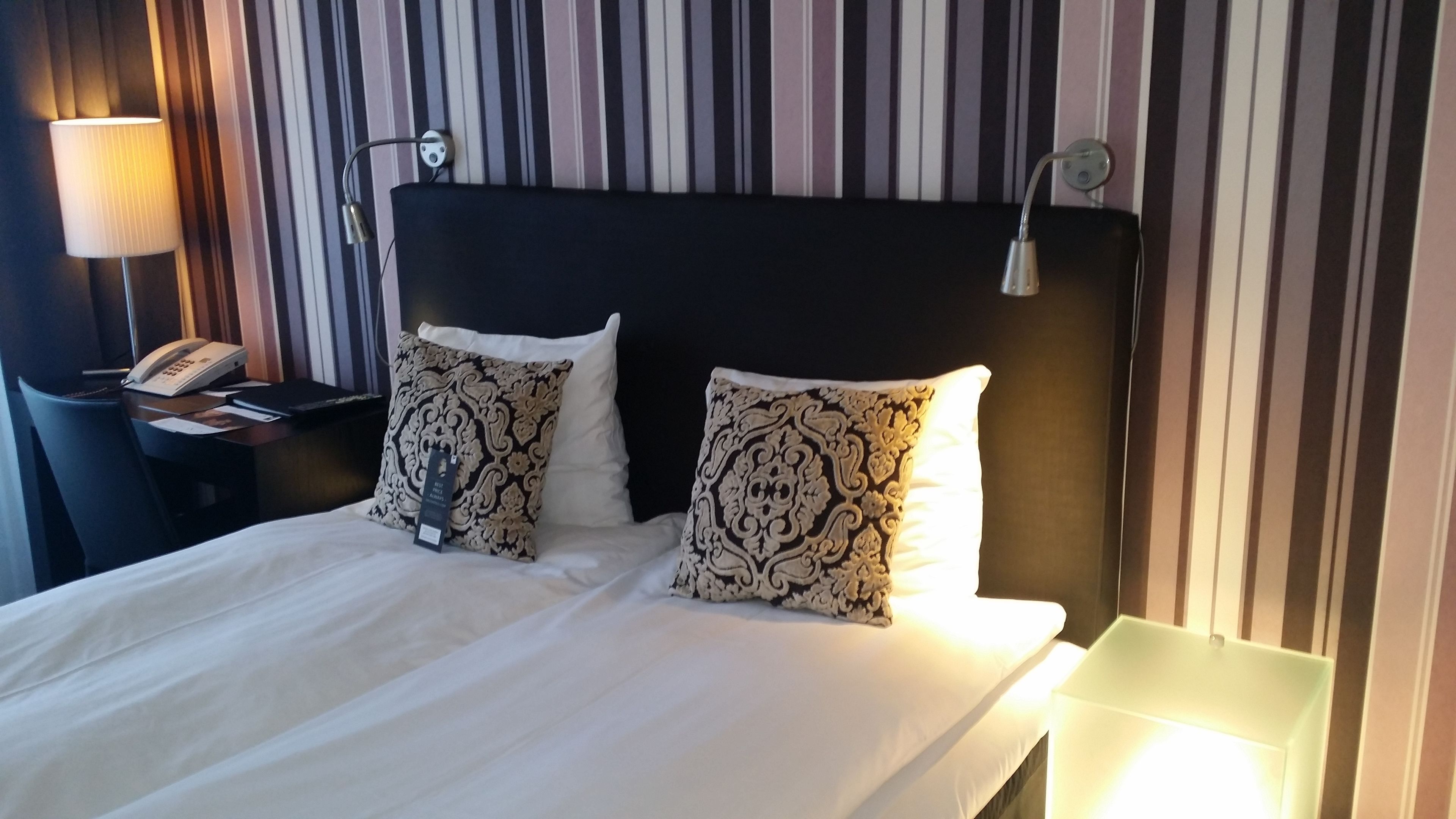 Habitación de hotel con una cama grande, dos almohadones decorativos, una lámpara de noche, un teléfono y un fondo de papel tapiz a rayas.