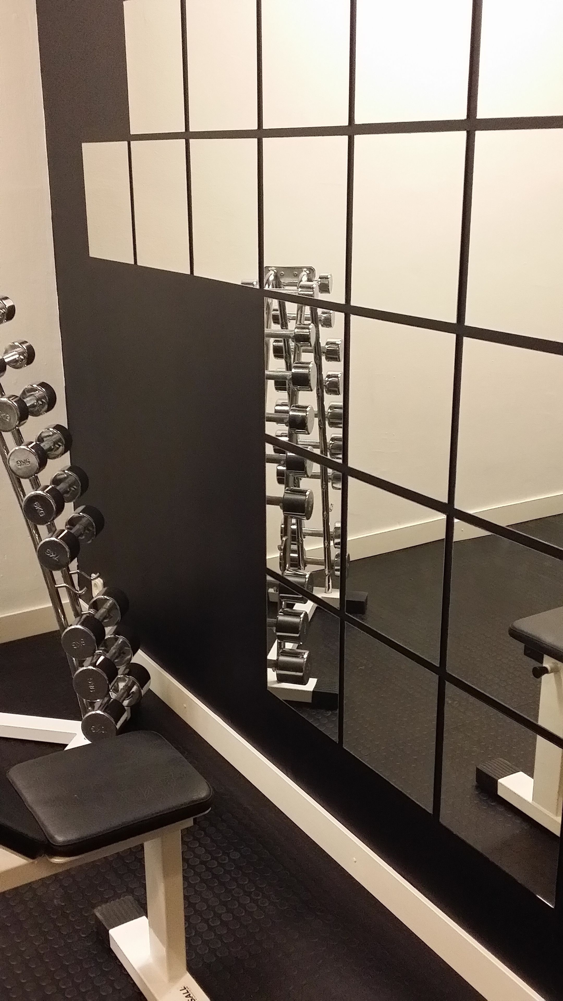 A gym with a mirrored wall, a black and white tiled wall, and a dumbbell rack.