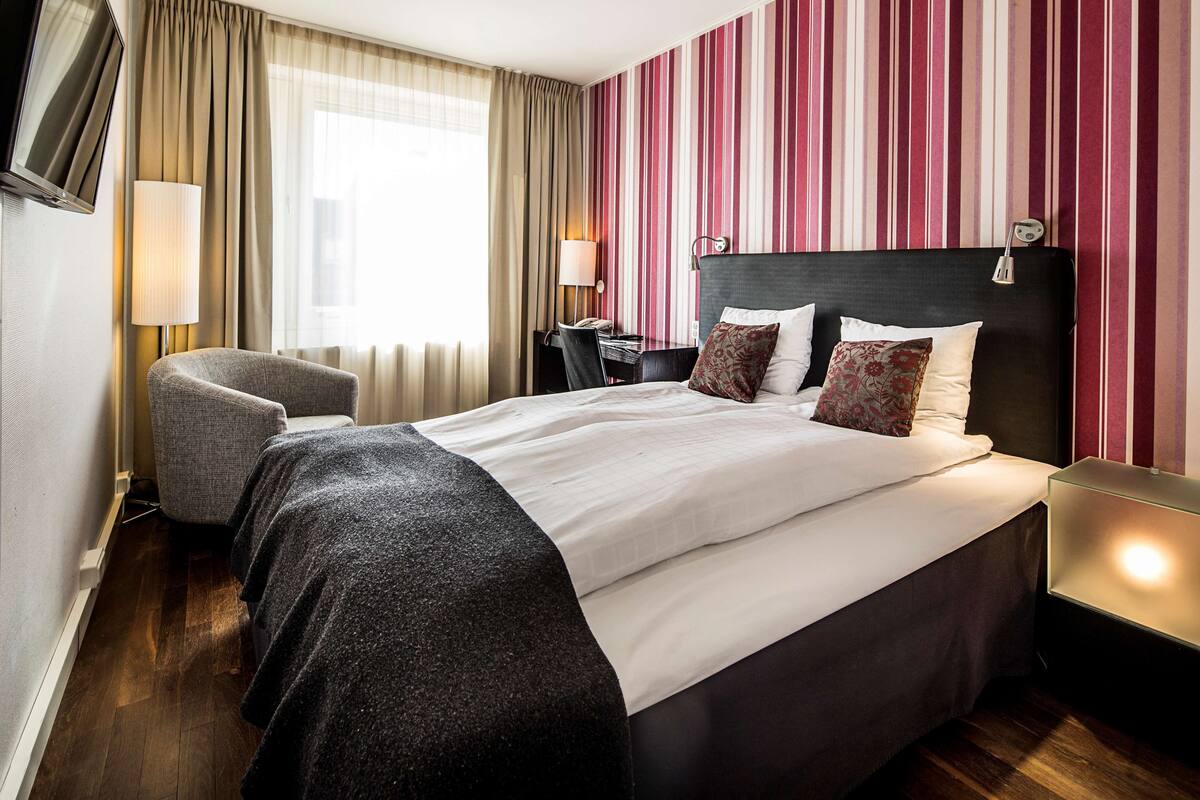 Habitación de hotel con una cama grande, un escritorio con lámpara, una silla y un televisor.