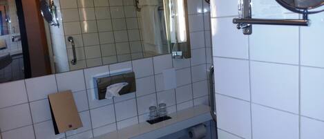 Un baño con espejo, dispensador de toallas, dispensador de jabón y porta papel higiénico.