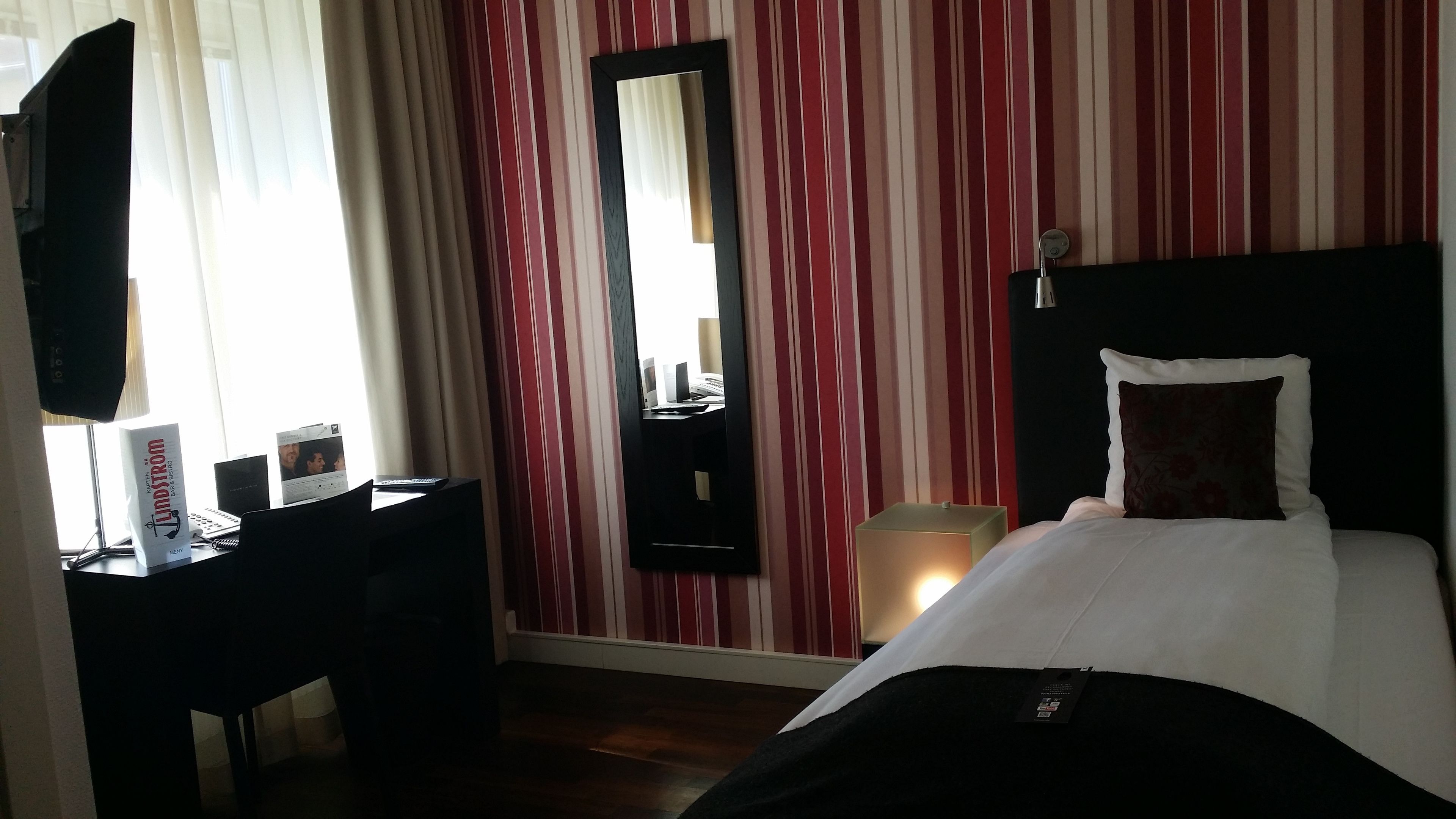 Habitación de hotel con cama, escritorio con computadora, espejo y papel tapiz a rayas.
