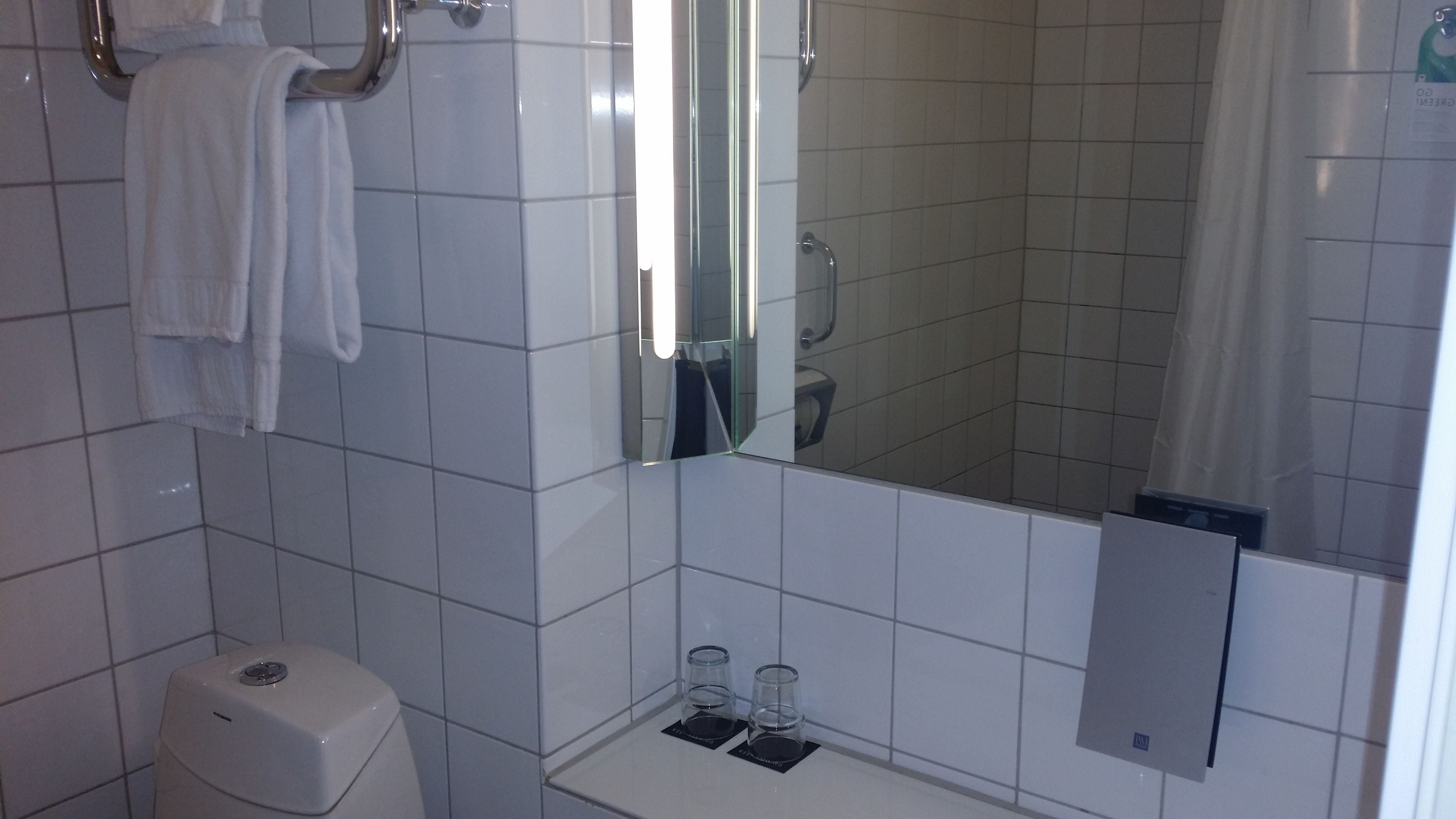 A bathroom with white tiles, a toilet, a sink with two glasses, and a mirror reflecting the toilet.