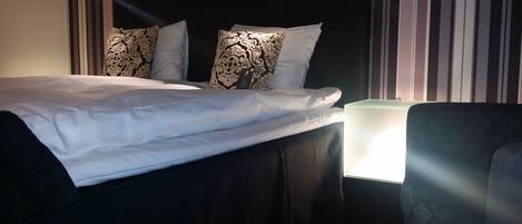 A neatly made bed with white linens and decorative pillows, a bedside lamp, and a chair.