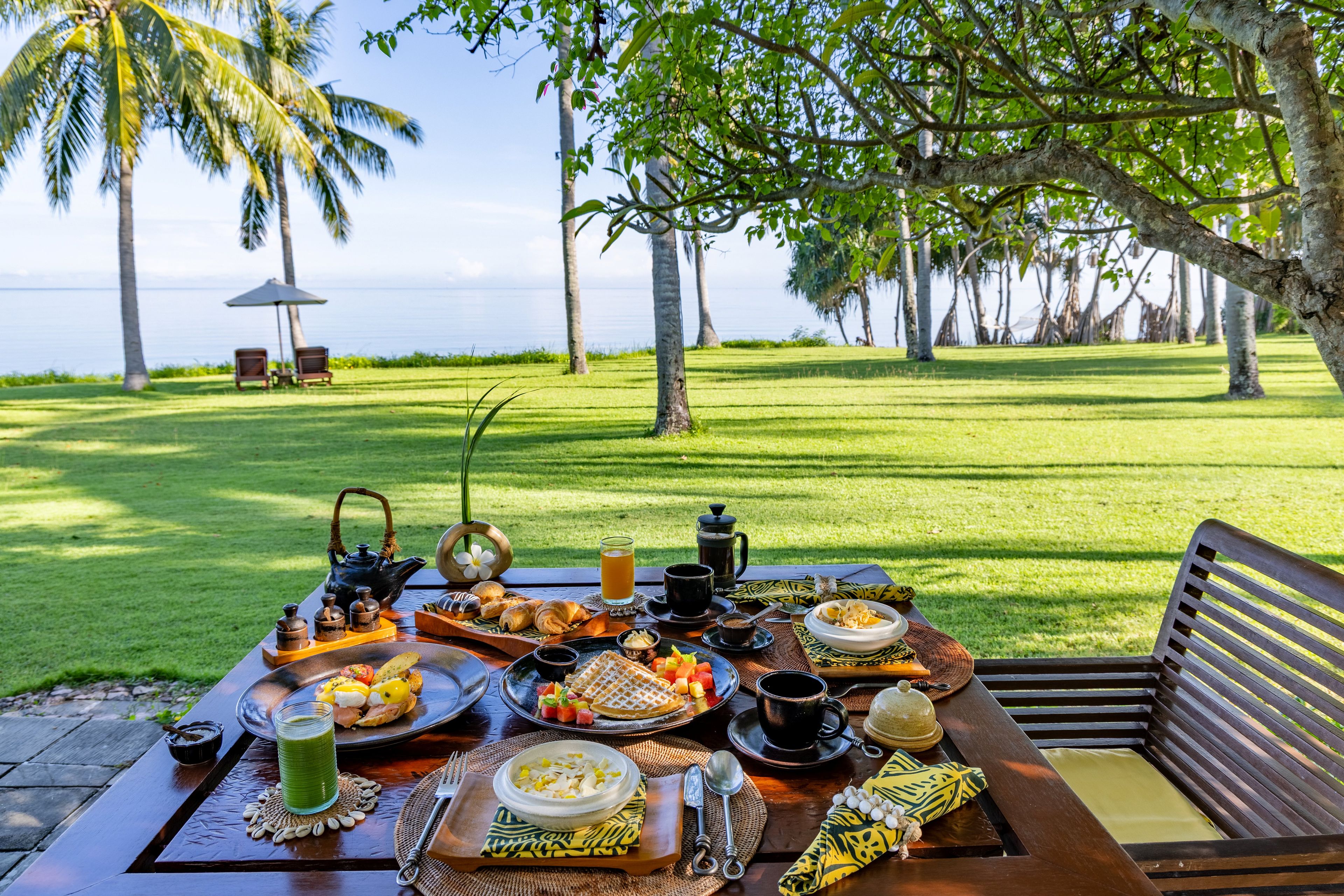 An outdoor dining area with a table set for breakfast, overlooking a grassy landscape with palm trees and a view of the sea.