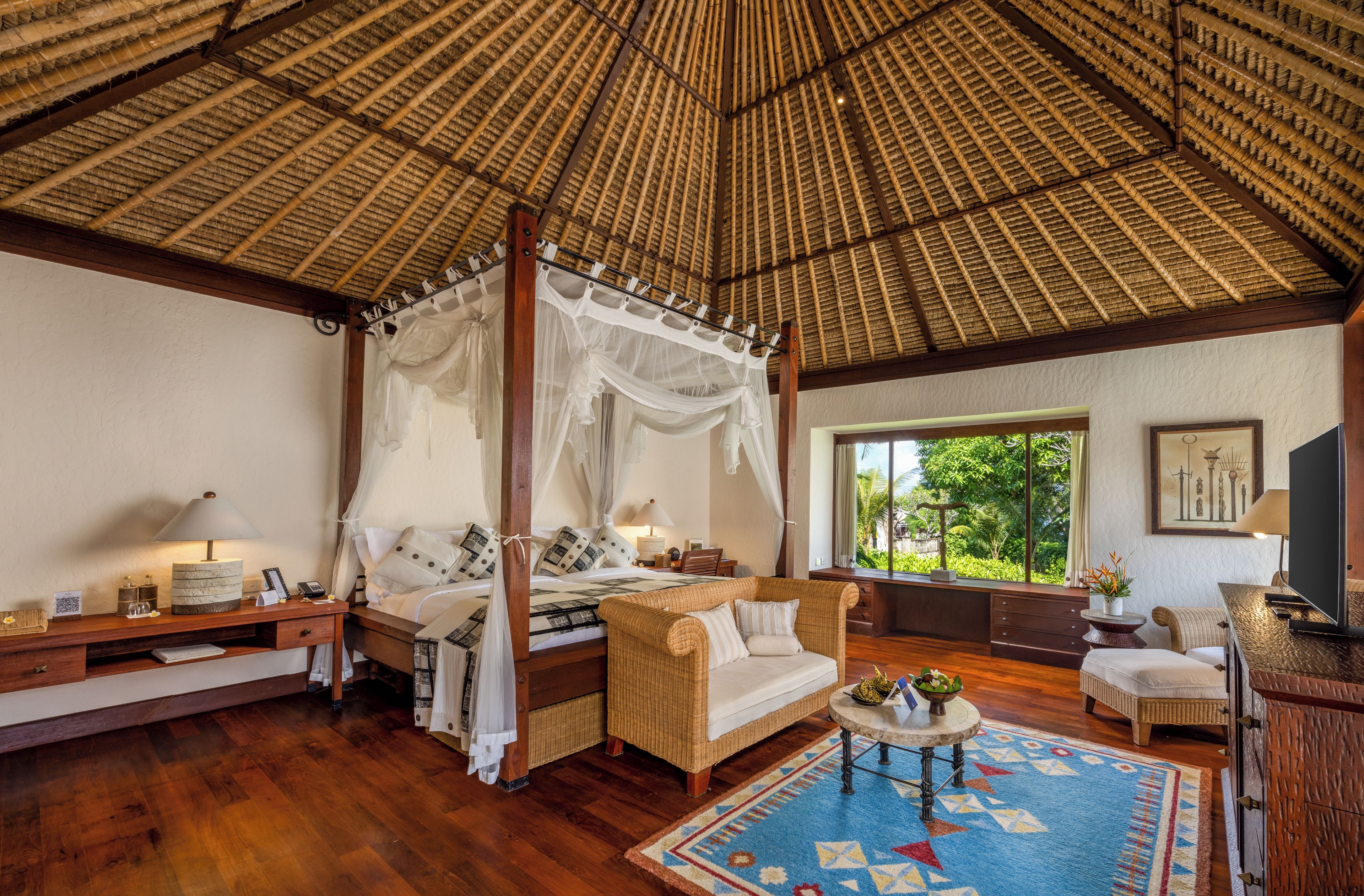 A room with a four-poster bed, a sofa, a dining table, and a large window with a view of greenery.