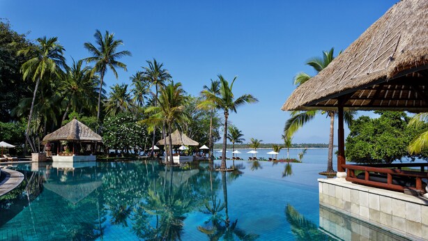 A clear blue infinity pool surrounded by palm trees and thatched-roof structures, with a view of the ocean. - The Oberoi, Lombok (Tanjung)