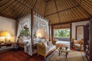 A four-poster bed with sheer curtains, a wooden floor, a wicker sofa, a small round table with a bowl, and a view of the outdoors through a glass door.