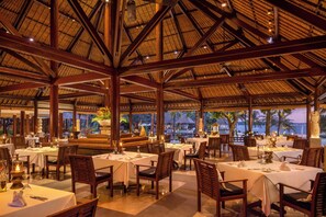 An indoor dining area with tables and chairs, a thatched roof, and a view of the sea.