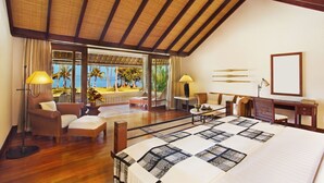 A spacious room with a large bed, a desk, and a chair. There is a window with a view of palm trees and the ocean. - The Oberoi, Lombok (Tanjung)