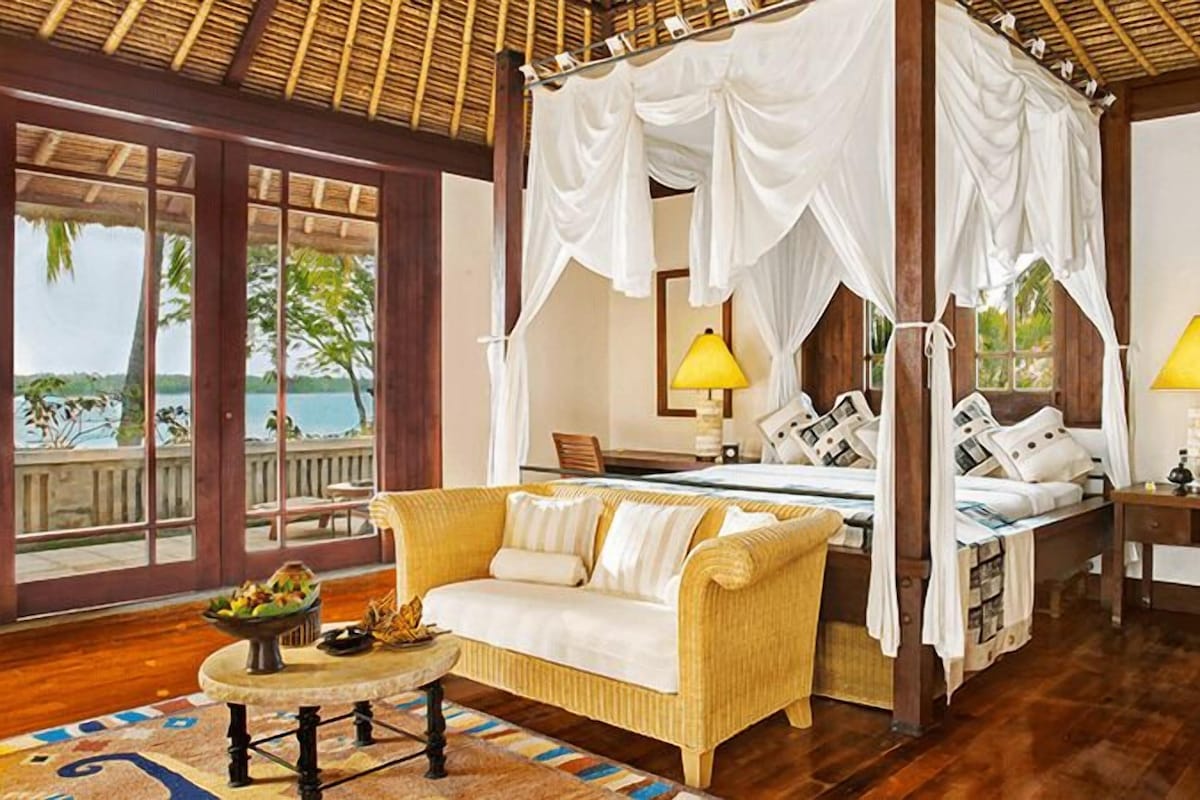 A four-poster bed with white drapes, a sofa, a bedside table, and a lamp.
