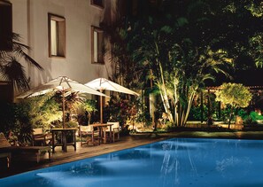 A nighttime pool area with umbrellas, lounge chairs, and a pool reflecting the surrounding trees.