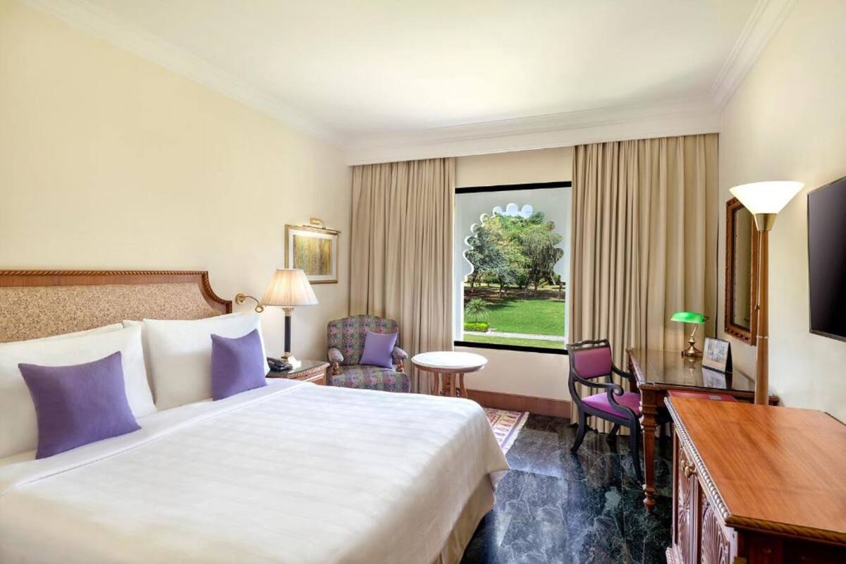 A hotel room with a large bed, a desk, a chair, a television, and a window with a view of greenery.