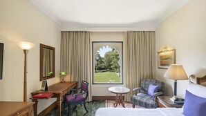 Ein Hotelzimmer mit einem Bett, einem Schreibtisch, zwei Stühlen, einem kleinen Tisch, einer Lampe, einem Gemälde und einem Fenster mit Blick auf einen Garten.