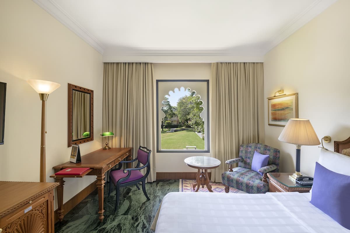 A hotel room with a bed, a desk, two chairs, a small table, a lamp, a painting, and a window with a view of a garden.